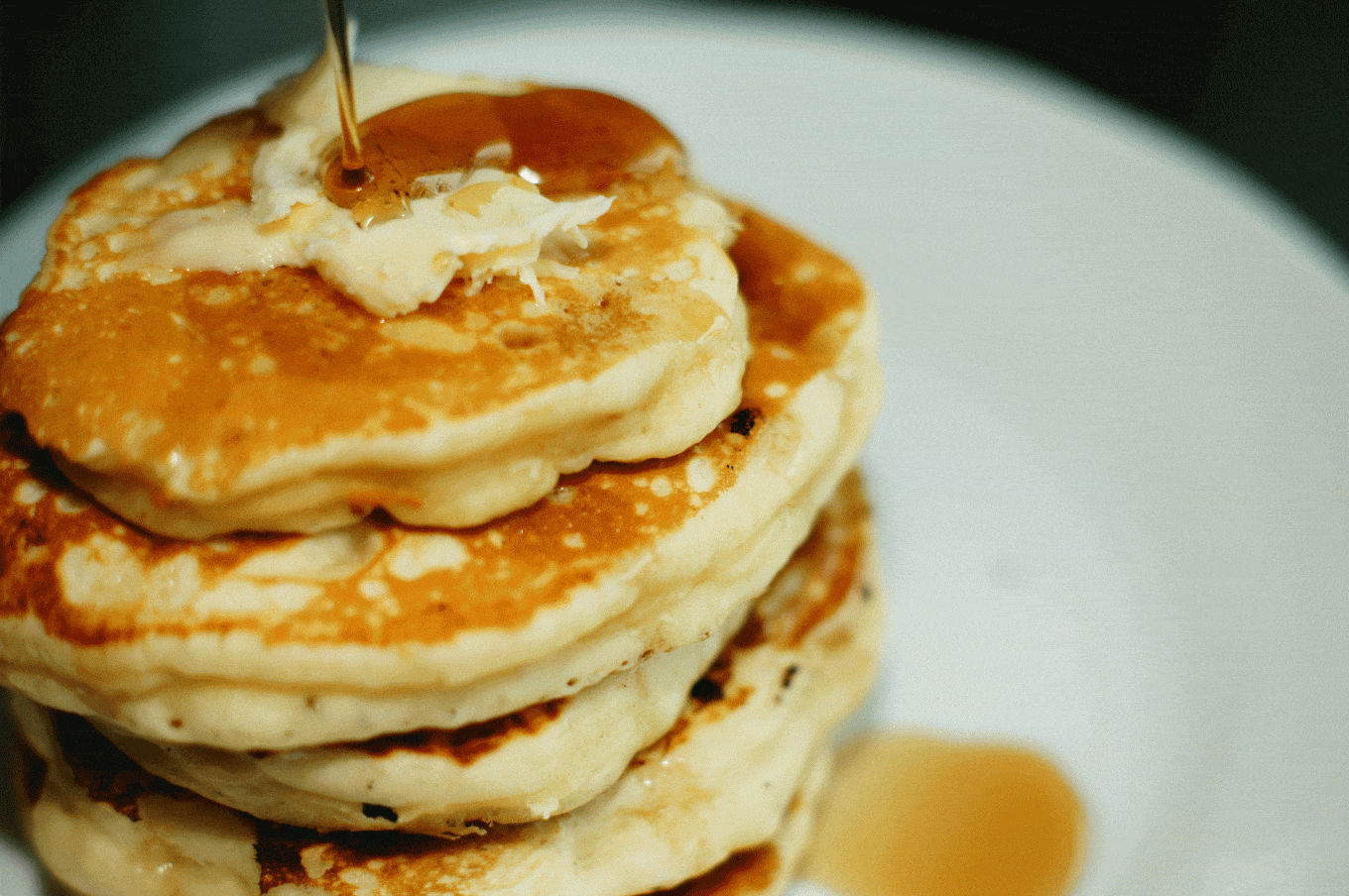 Golden maple syrup being drizzled on a pat of butter on top a stack of fluffy pancakes.