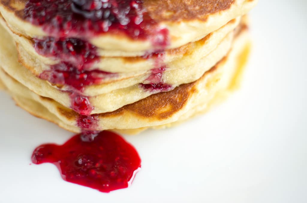 A drizzle of mixed berry compote falls down the side of a stack of delicious and fluffy lemon mascarpone pancakes, served up on a white plate for breakfast/brunch.