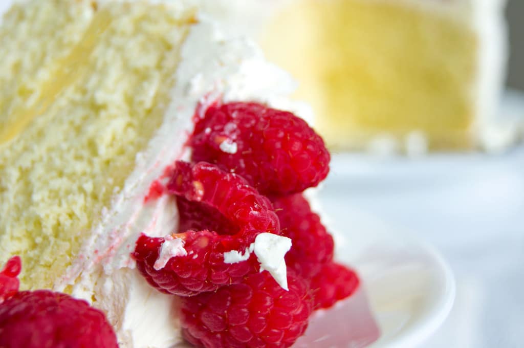 A slice of lemon curd cake on its side with white chocolate swiss buttercream frosting and raspberries on top.