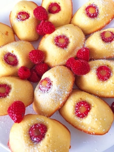 An overhead shot of a batch of Lemon-Raspberry madeleines on a white plate with raspberries.