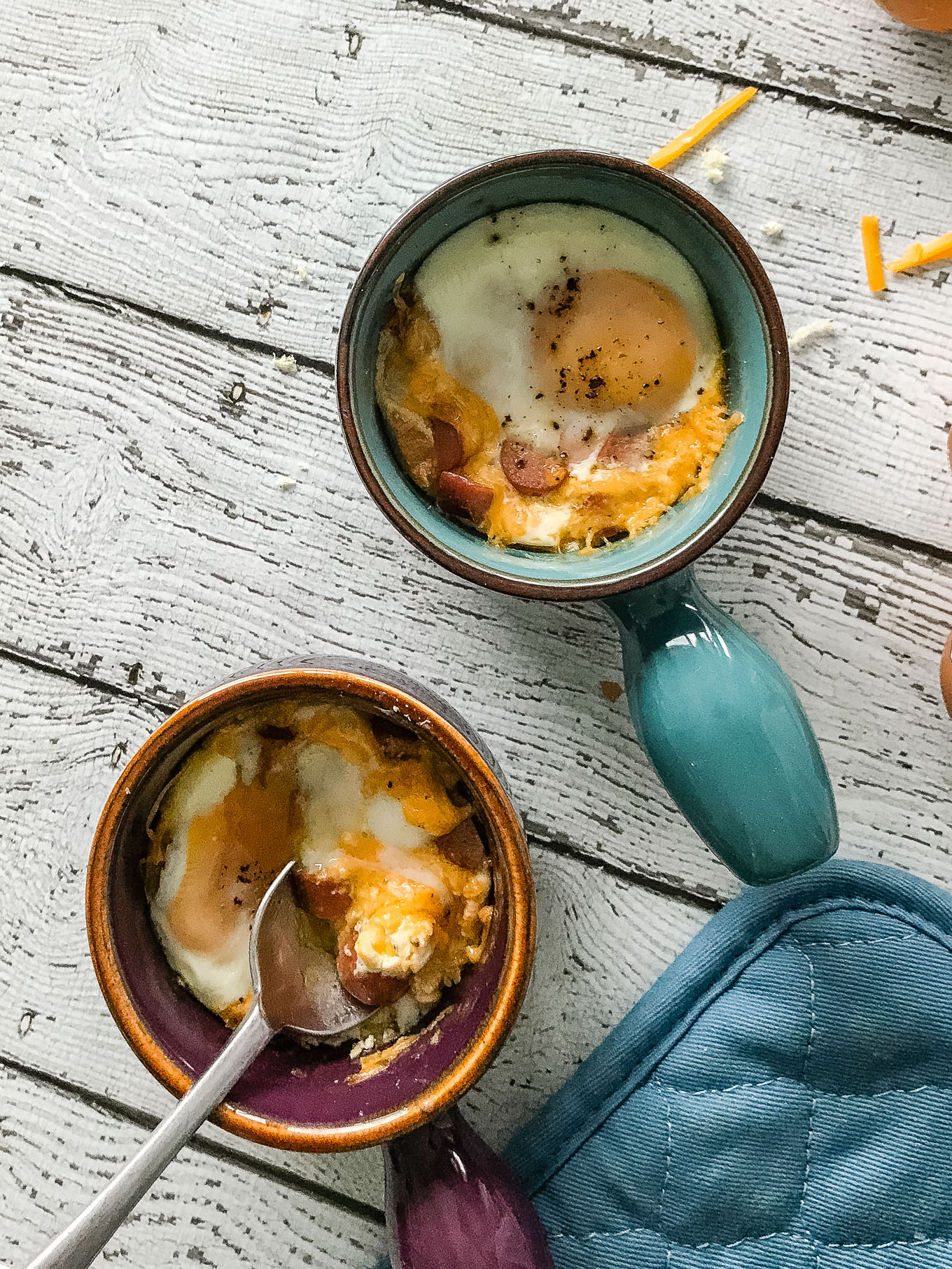 A vertical overhead shot of Cheesy Oeufs en Cocotte / Eggs in Pots, with a spoon cutting into one of the eggs. 