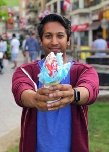 A photo of Riz wearing a maroon hoodie, holding forward a colourful ice cream cone.