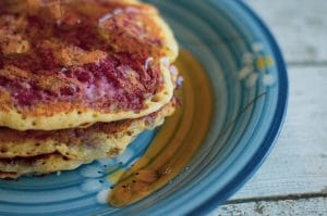 A vibrant photo with a stack of raspberry swirl pancakes on the left side of the photo, plated on a deep blue plate with overflowing maple syrup.