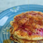 A photo of maple syrup being poured on to a stack of raspberry swirl pancakes.