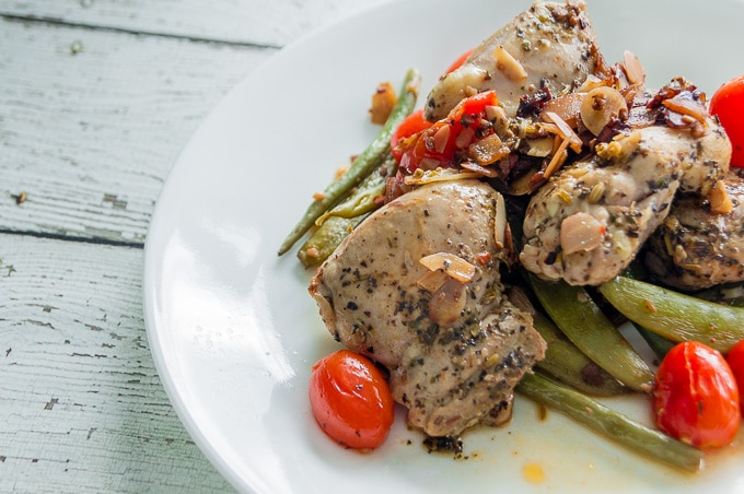 A plate of chicken, green beans, sugar snap peas, and cherry tomatoes on a white wooden table.