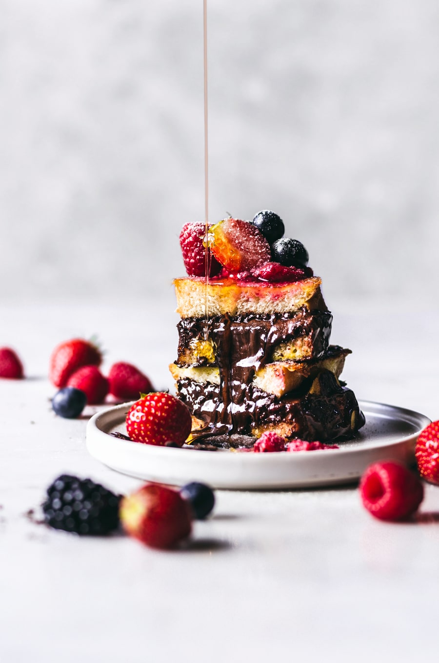 Maple syrup being drizzled on to stacked Nutella & Dark Chocolate Stuffed French Toast, with some berries scattered around it.