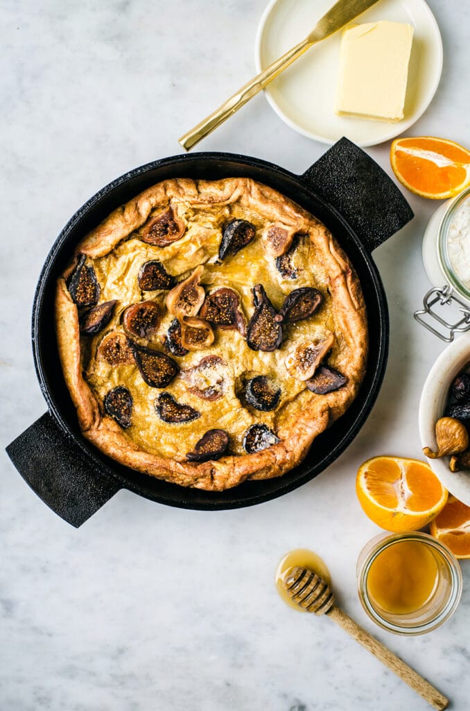 A flatlay of a Dutch Baby Pancake with figs on a marble top with butter, oranges, and honey surrounding it.