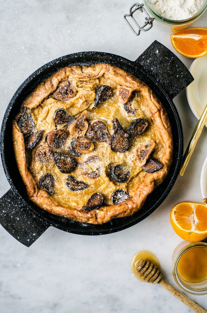 An overhead photo of the fig Dutch Baby Pancake with honey, sliced oranges, and flour in the background.
