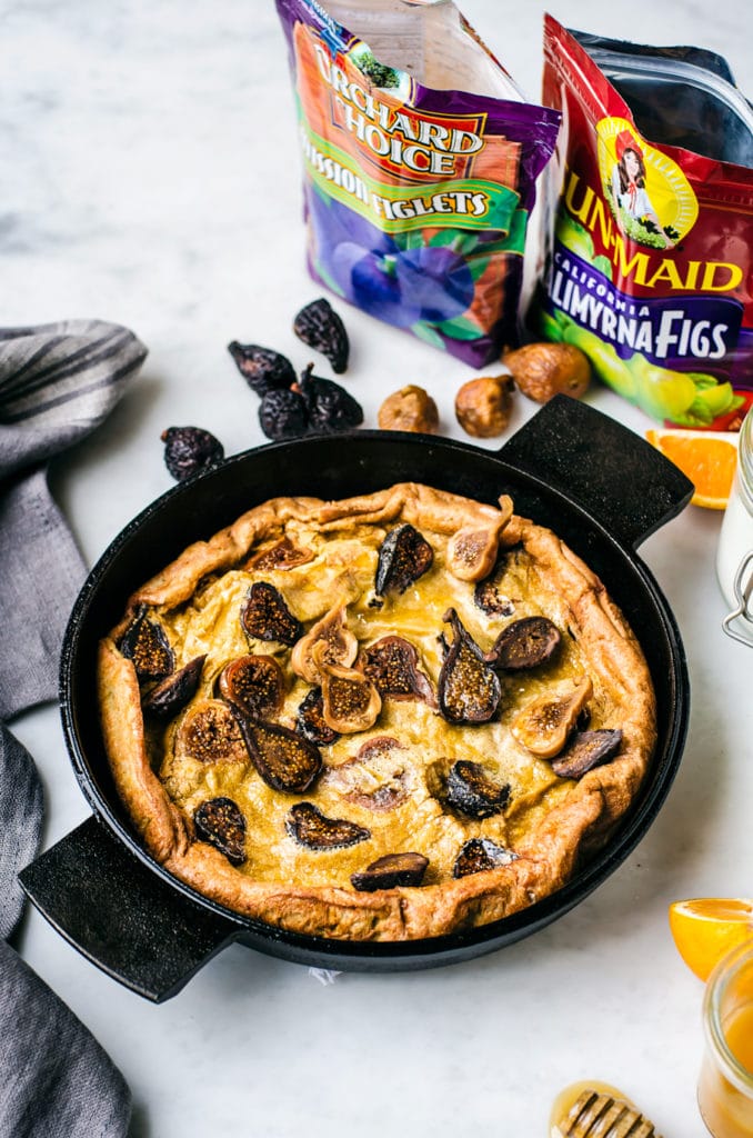 A fig Dutch Baby Pancake in a cast iron pan with Valley Fig Growers' products (Orchard Choice Mission Figlets, and Sun-Maid Calimyrna Figs) in the background.