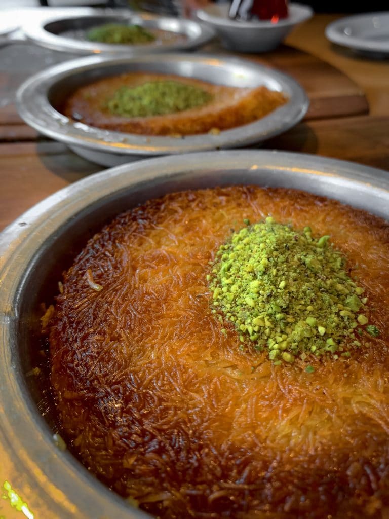 3 silver trays of kunafa (decorated with pistachios) in a row atop a wooden table.