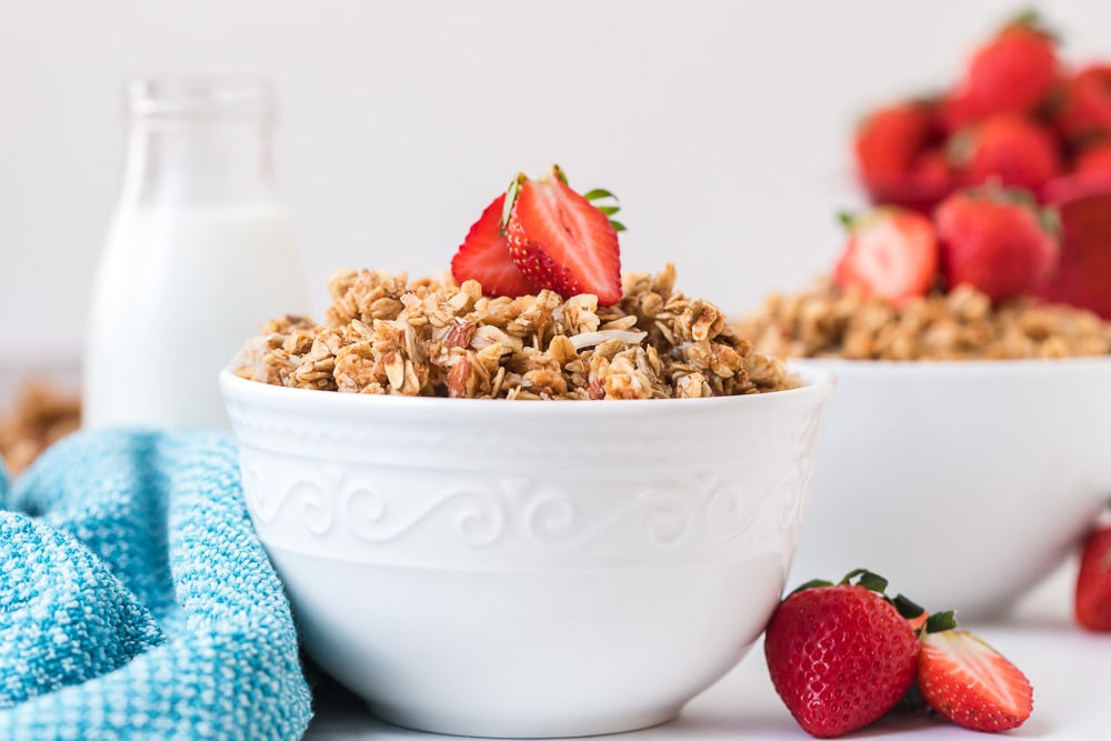 Two bowls of granola with sliced strawberries, and a bottle of milk in the background.