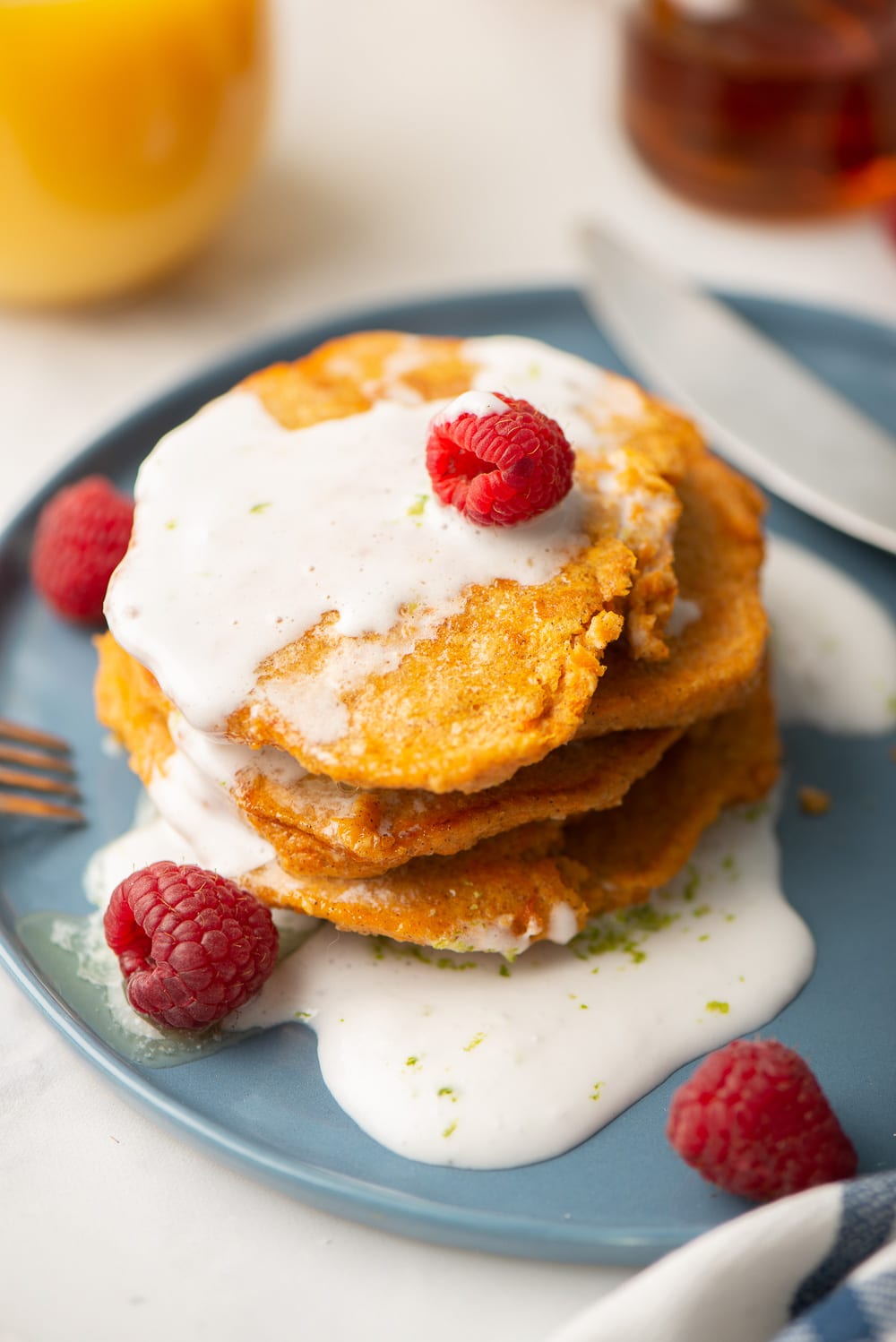 A close-up of a stack of gluten-free sweet potato pancakes topped with coconut-lime whipped cream and some raspberries on a blue plate.