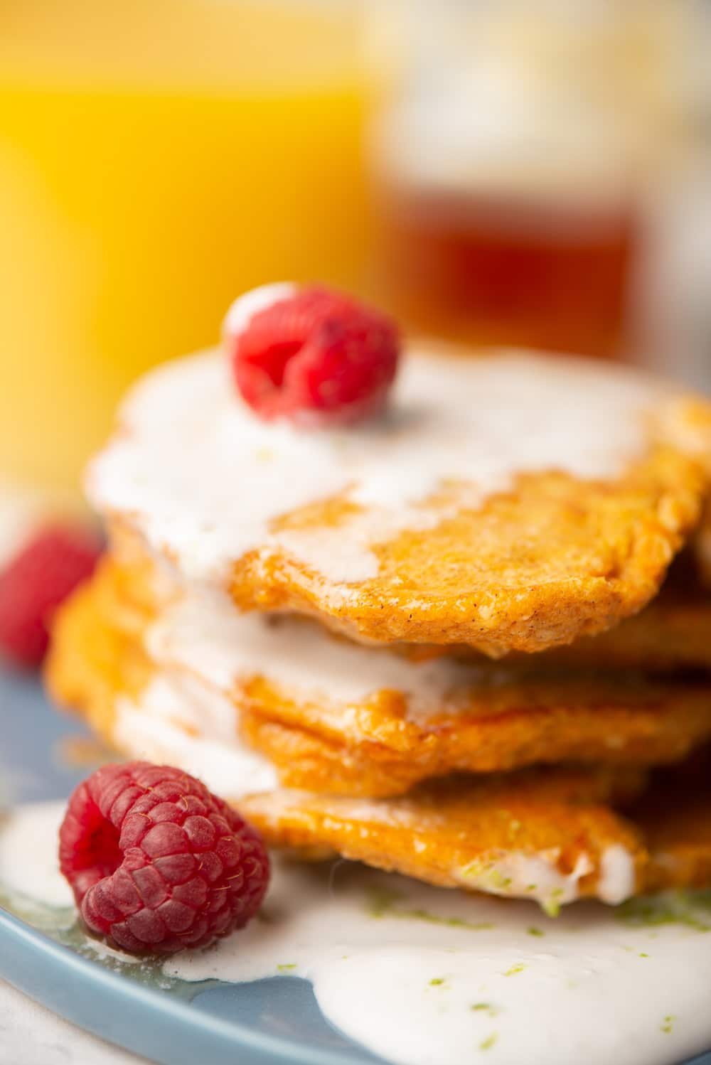A close-up of a stack of fluffy sweet potato pancakes that have been topped with a lime-infused coconut cream and raspberries. The background has been blurred out.