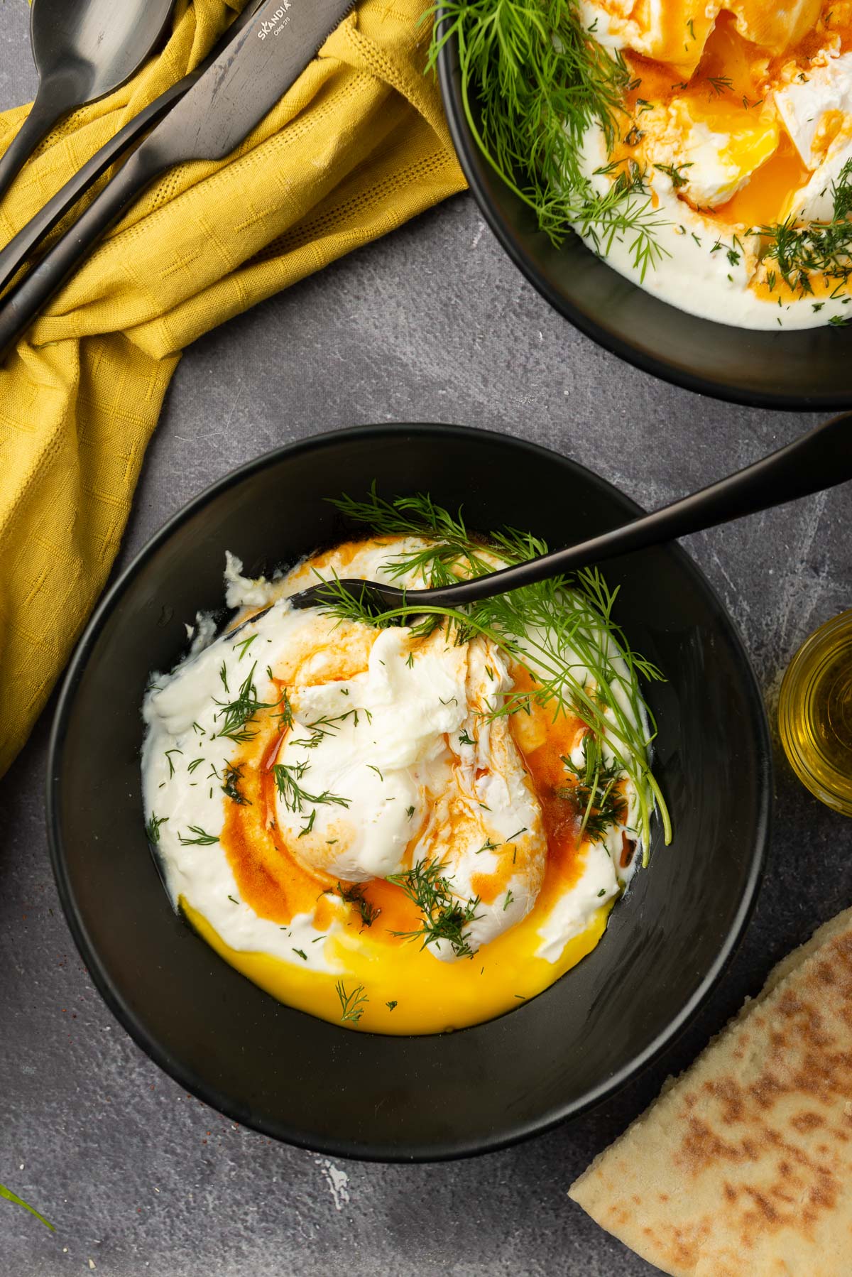 An overhead shot of Turkish eggs in yoghurt, served in a black bowl with dill and a popped yolk.