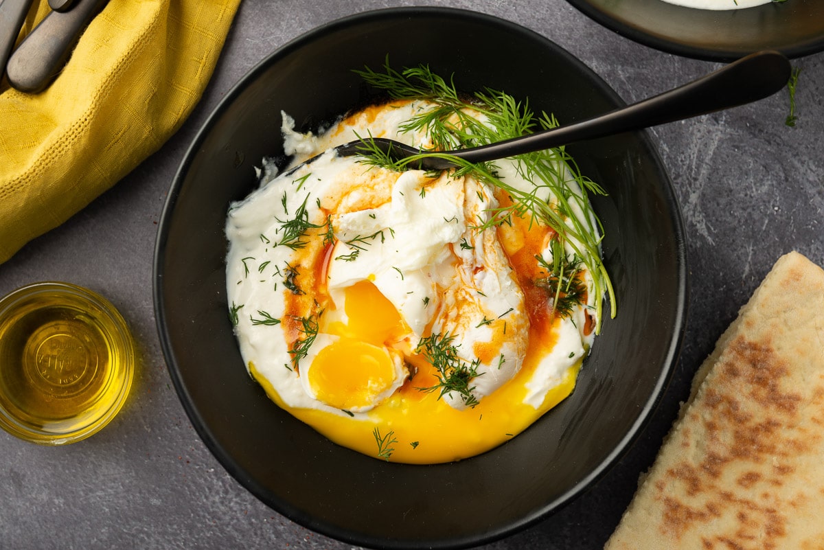A flatlay of Turkish eggs in a black bowl. A black spoon has cut open one of the poached eggs so that the yolk has oozed out to the spiced yogurt.