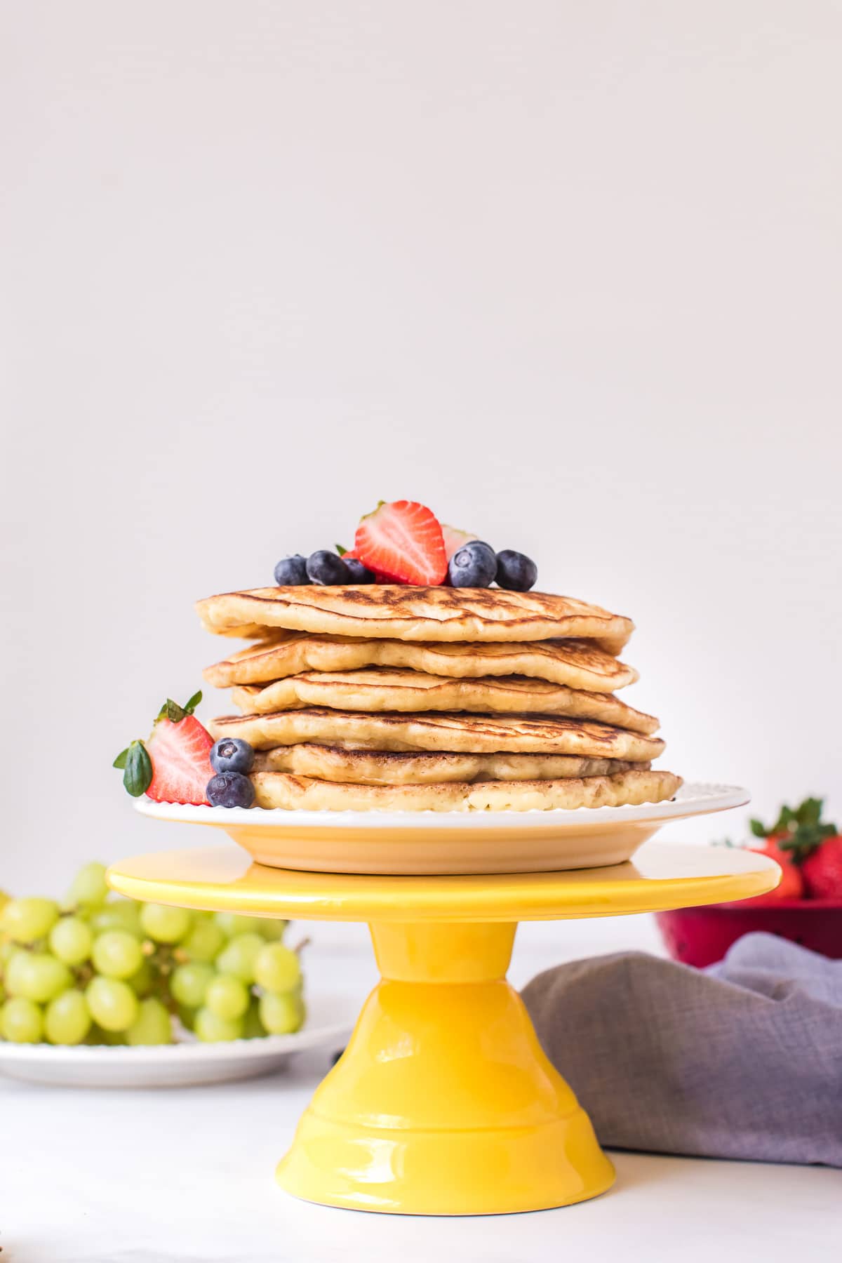 A tall stack of easy pancakes on a white plate set atop a yellow cake stand, and garnished with berries.