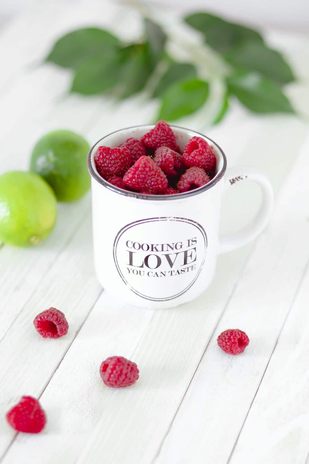 A white mug filled with raspberries. The mug says "Cooking is love you can taste"