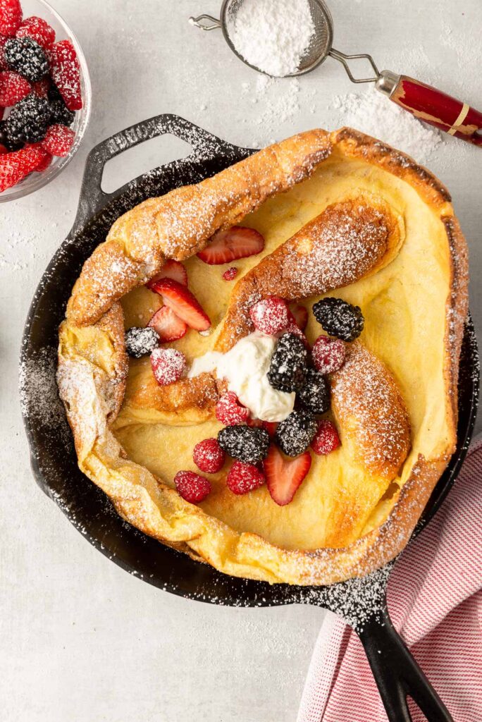 A portrait photo of a Dutch baby pancake in a black cast iron pan, decorated with strawberries and blackberries.