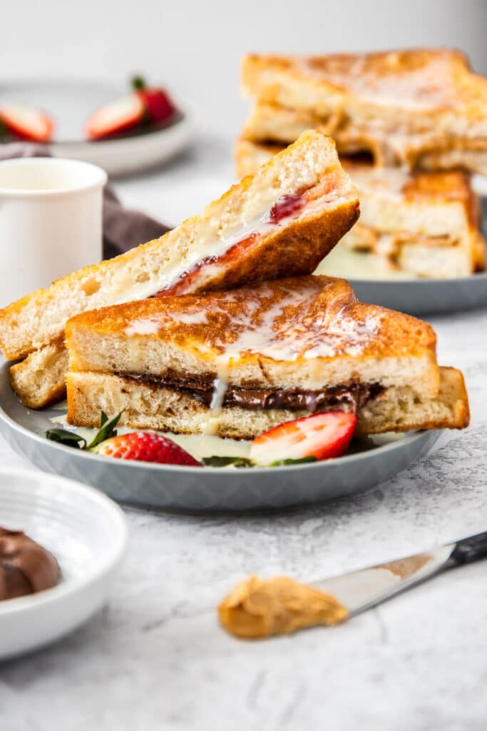 2 Hong Kong style French Toast sandwiches, one is stuffed with strawberry jam, while the other is filled with Nutella. In the distance a third sandwich, stuffed with peanut butter is visible.