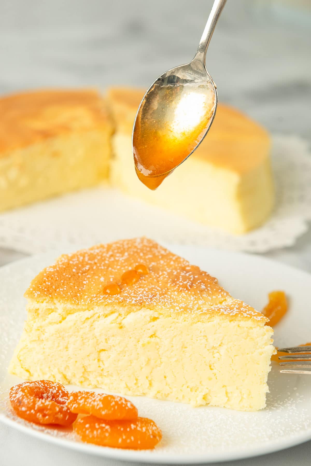 A slice of Japanese cheesecake on a white plate; a spoon above it is dripping apricot glaze on to it.
