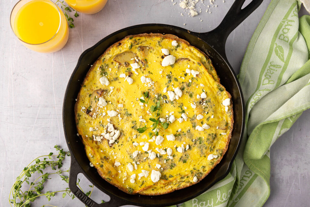 An overhead view of a cooked potato frittata, garnished with thyme and goat's cheese in a cast iron pan.