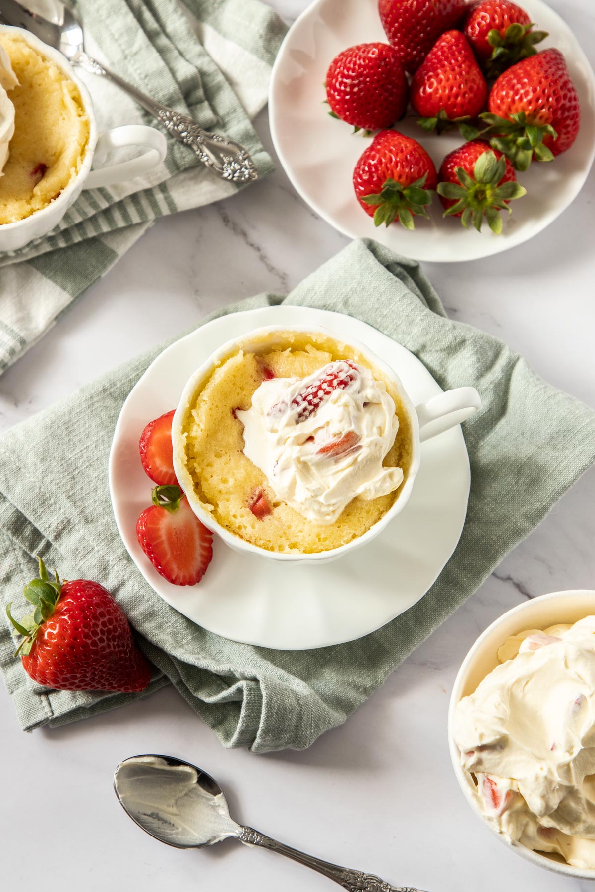 Top down photo of strawberry shortcake in a mug surrounded by fresh strawberries, cream, and table linen.