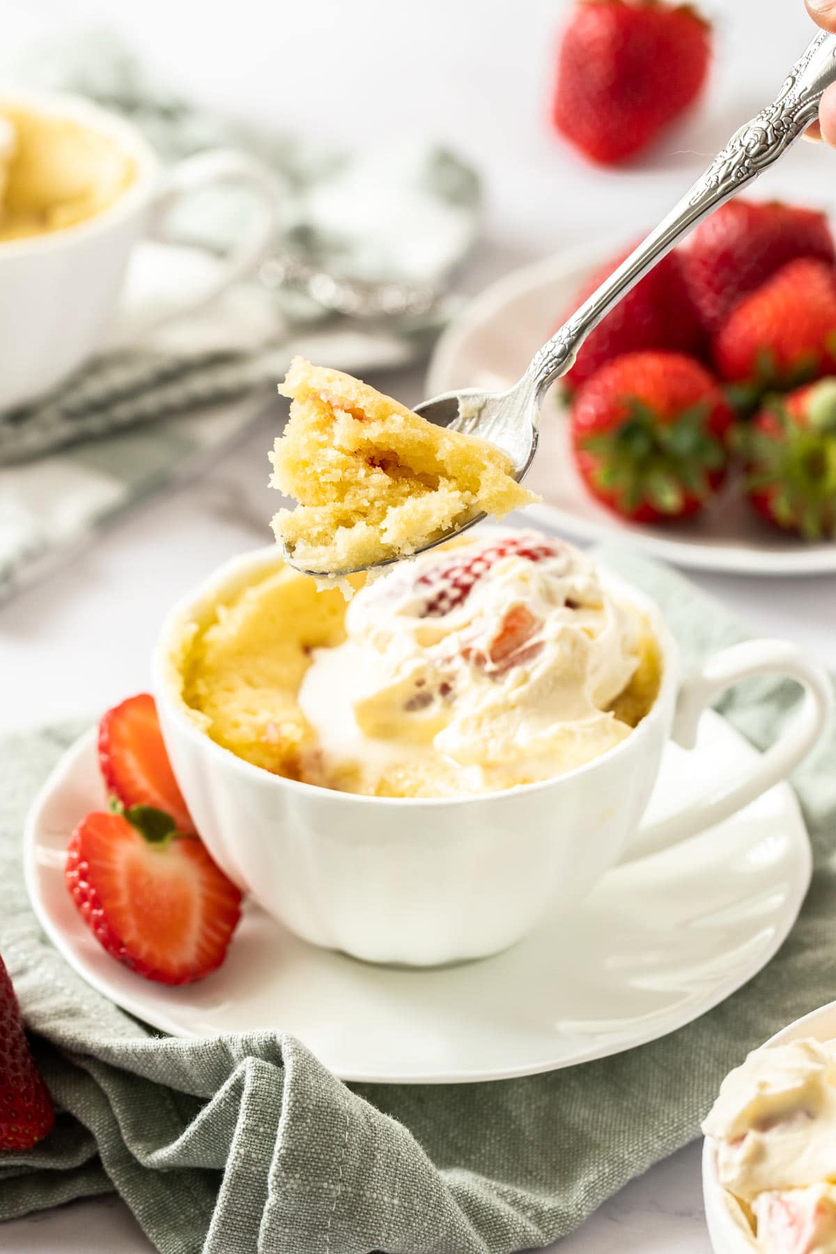 A spoonful of strawberry shortcake in a mug with the mug cake in the background.