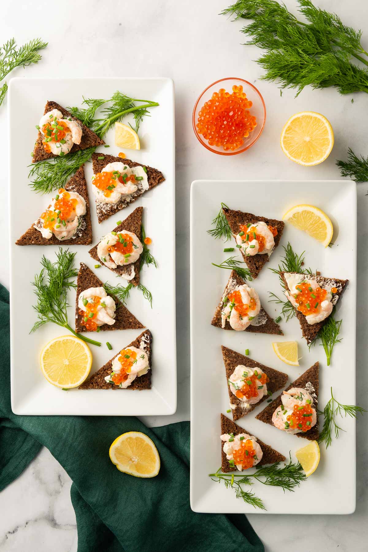 An overhead shot of two rectangular platters of Swedish Toast Skagen decorated with dill, lemon, and a green napkin in the background.