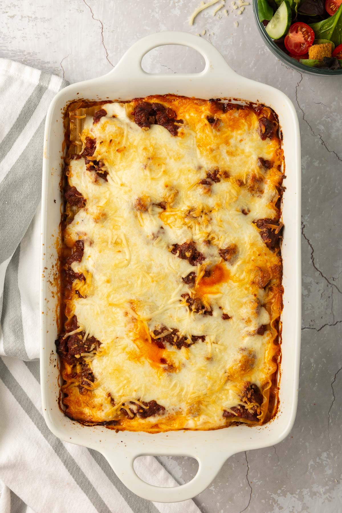 A fully cooked lasagna in a white dish accented with a white and grey kitchen towel.