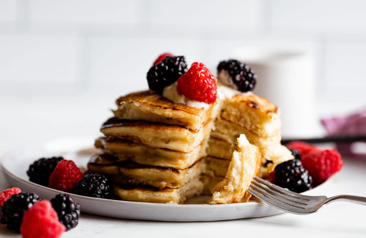 A bite of yogurt pancakes on a fork, placed next to a stack of partially eaten fluffy pancakes.