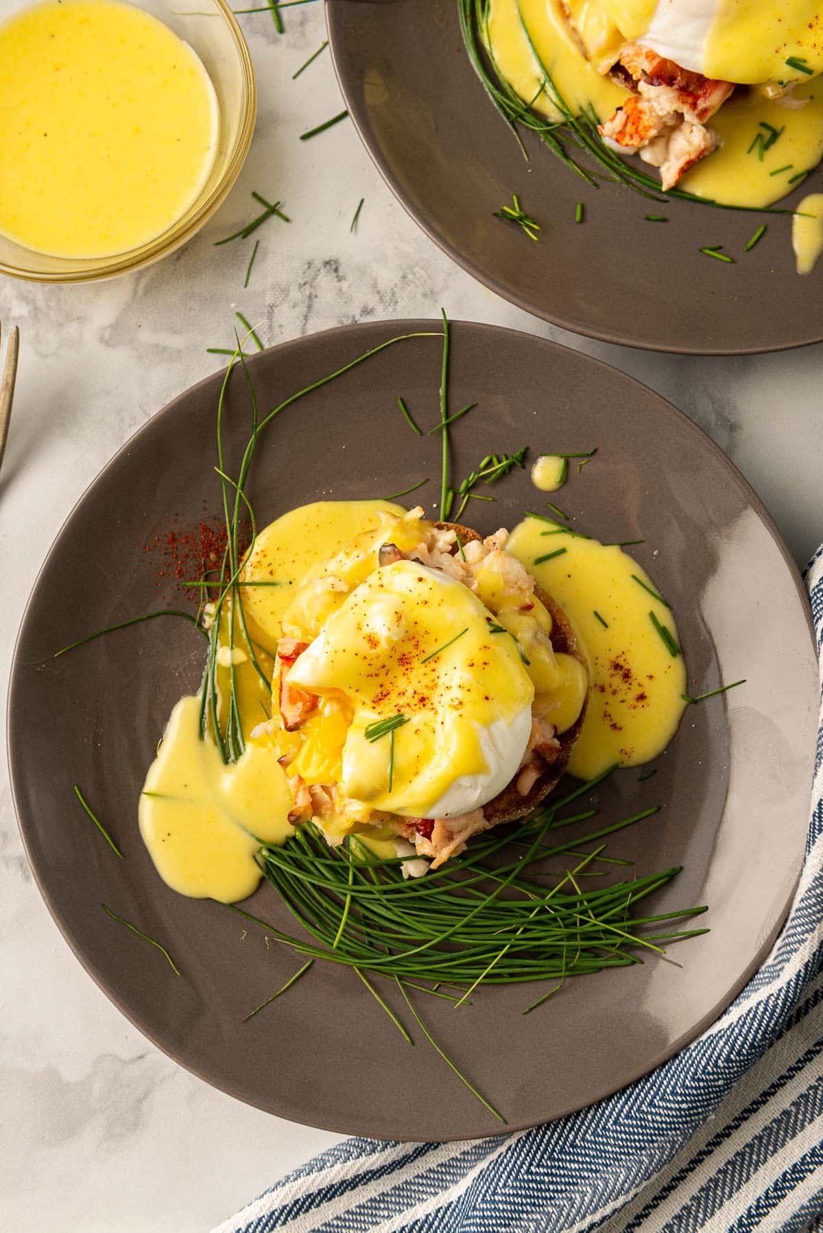 Yolk drizzling down the side of the lobster Benedict, decorated with chives on a dark plate.