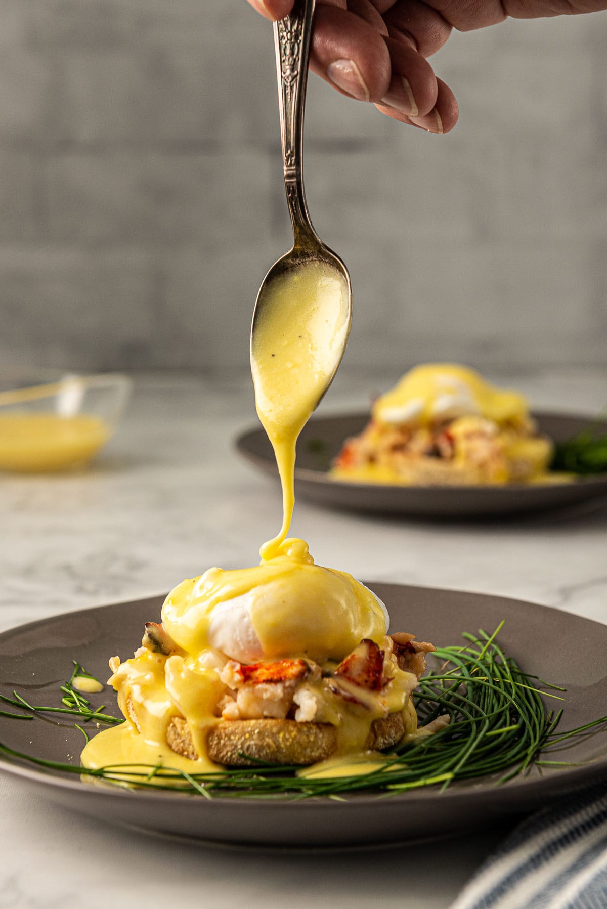 Hollandaise being drizzled off a spoon on to the top of a poached egg Benedict with lobster meat.
