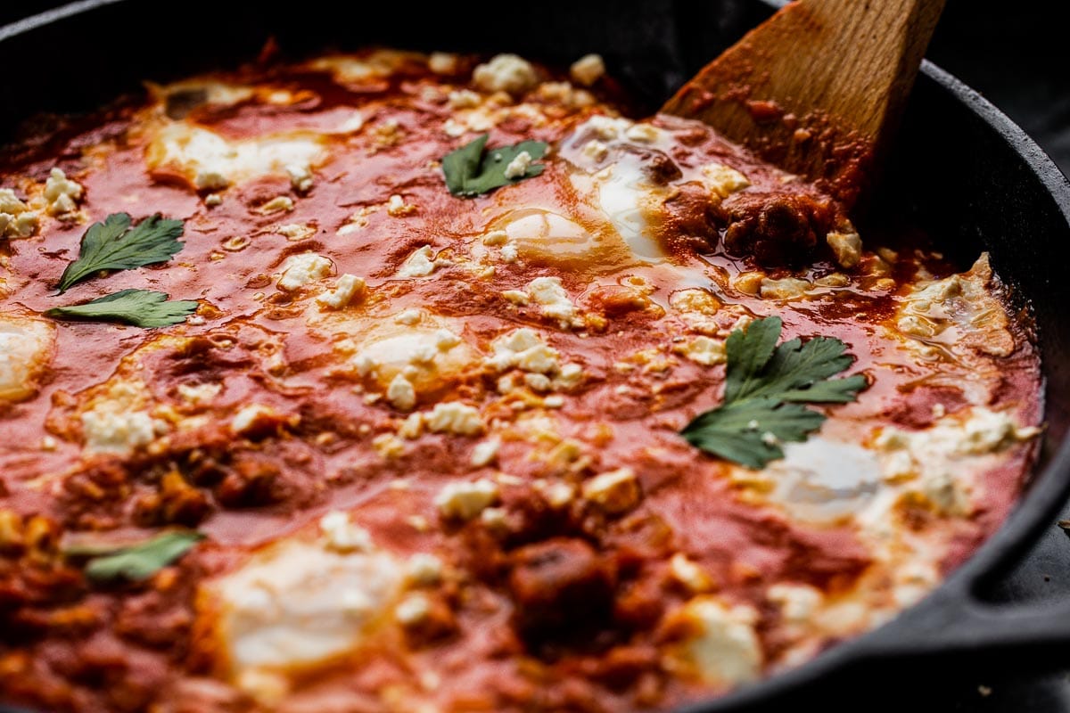 A wooden spoon digging into shakshuka to reveal crumbled merguez pieces.