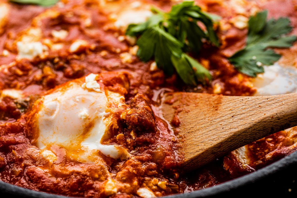 Wooden spoon lifting a poached egg from a skillet of merguez shakshuka.