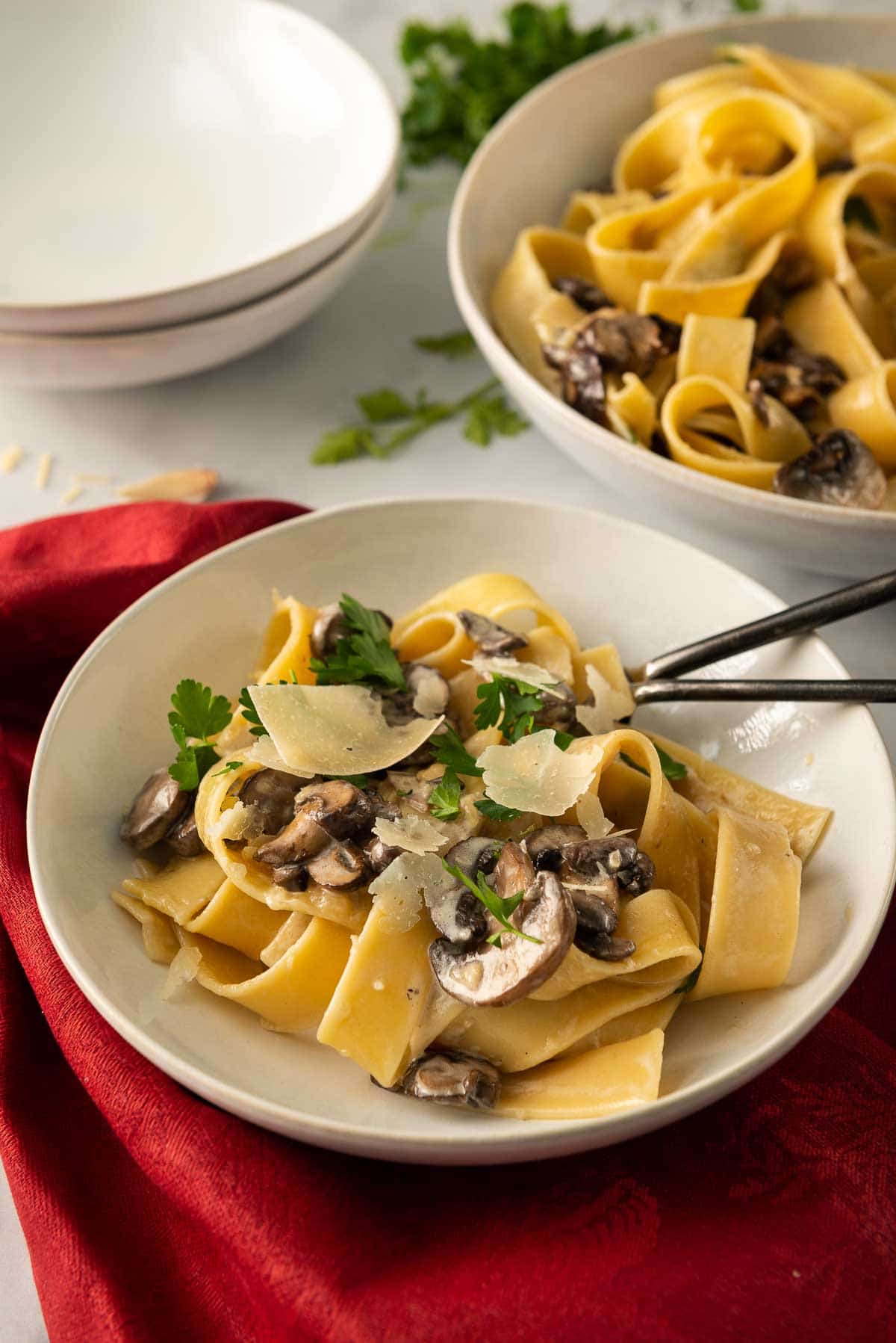 A plate of creamy mushroom pappardelle pasta that's ready to eat next to a serving bowl and some empty pasta bowls.