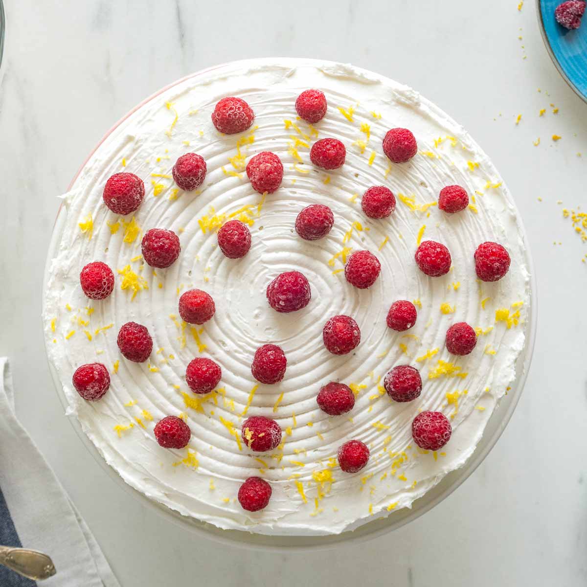 A top down photo of a white cake with lemon zest and raspberries on top. 