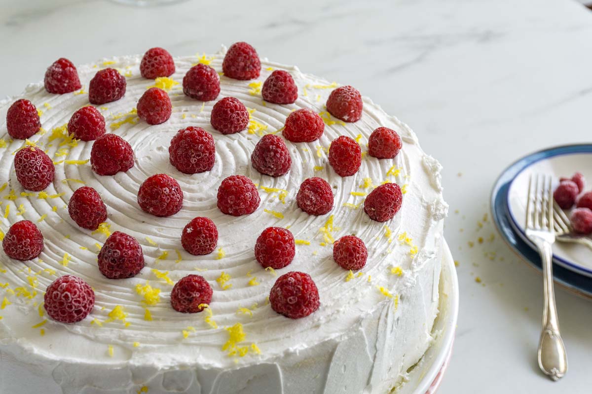 A lemon cake with white chocolate buttercream frosting, raspberries, and lemon zest.
