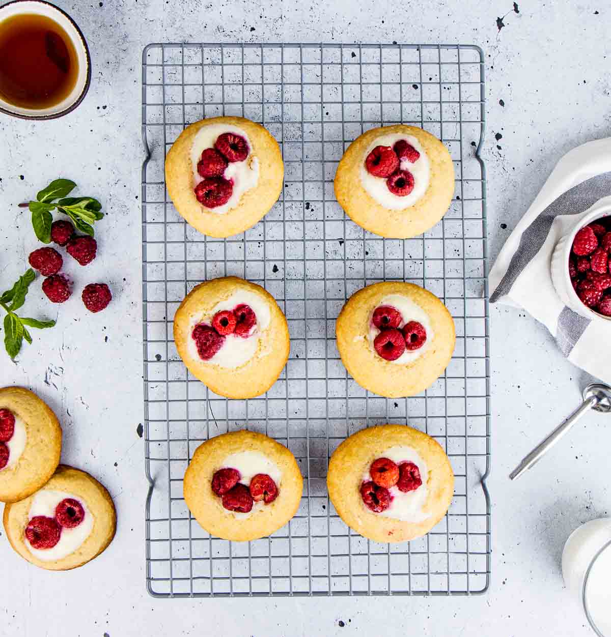 Six raspberry cheesecake cookies on a wire rack, with two more thumbprint cookies on the side.