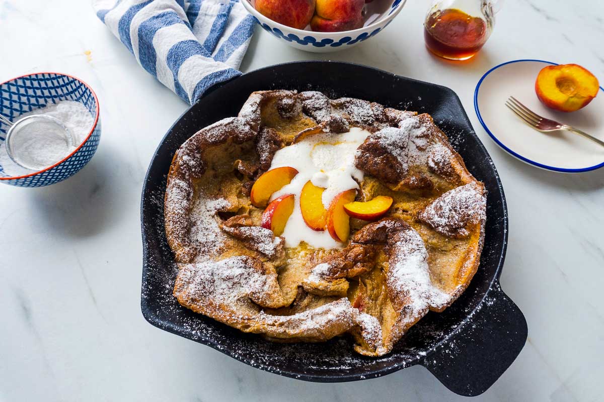 A peach Dutch Baby Pancake with fresh peaches, maple syrup, and icing sugar around it.
