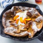 A Peach Dutch Baby Pancake in a cast iron pan. It has been garnished with fresh peaches, icing sugar, and melted ice cream.