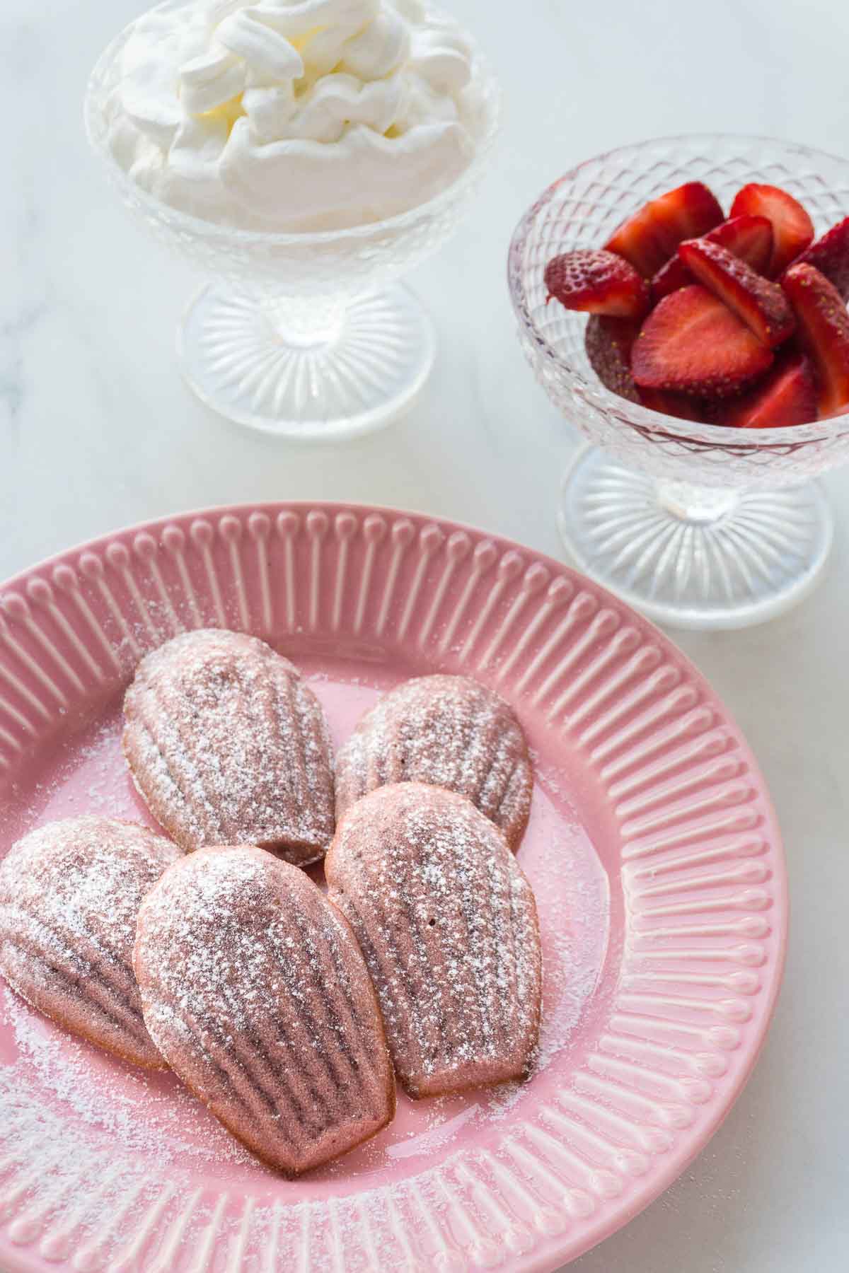 Pink strawberry madeleines in front of two bowls - one with strawberries, the other with cream.