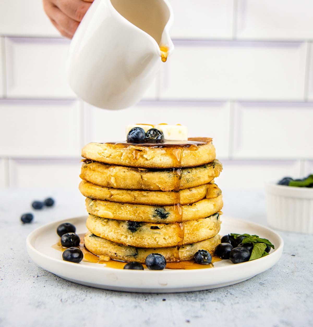 Maple syrup runs down the side of a stack of 6 fluffy blueberry pancakes.