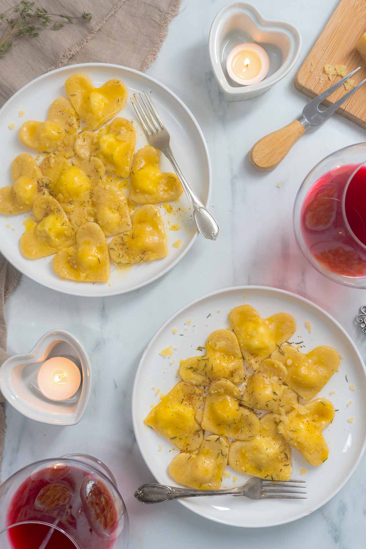 Two plates of ravioli hearts on a white table with a heart shaped candle next to them.