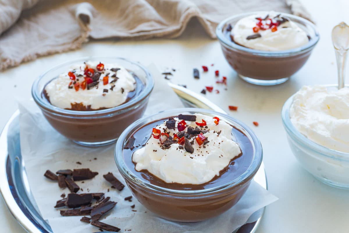 Three chocolate puddings - two in the front, and one in the background next to a bowl of whipped cream.