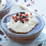 A close-up photo of a glass ramekin filled with dark chocolate pudding. There is a dollop of whipped cream with Espelette pepper and chocolate chunks.