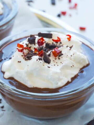 A close-up photo of a glass ramekin filled with dark chocolate pudding. There is a dollop of whipped cream with Espelette pepper and chocolate chunks.