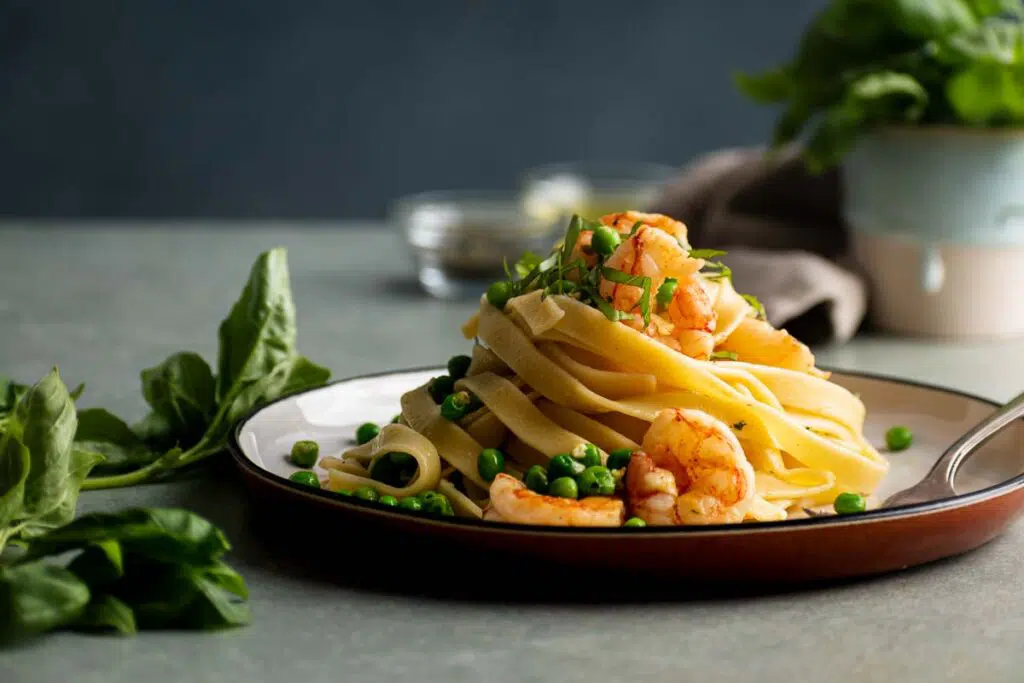 A plate of fettuccine pasta garnished with shrimp, peas, and finely chopped herbs, set on a dark ceramic plate. Fresh spinach leaves are scattered on the table, and a blurred backdrop shows additional ingredients, all contributing to a deliciously appetizing scene.