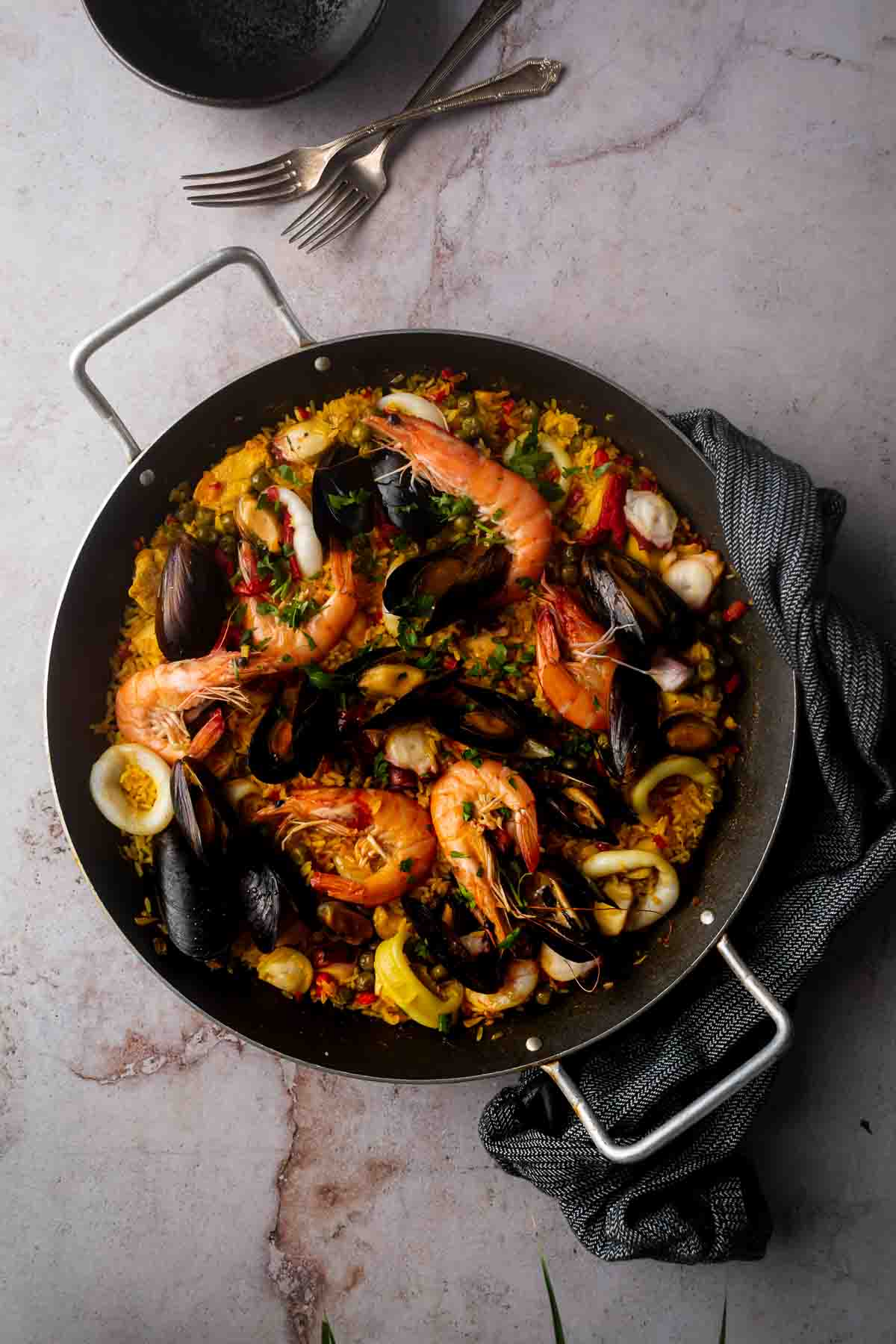 A top-down photo of cooked paella Valenciana with seafood in a paellera. A black hand towel accents the photo.