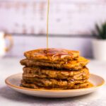 A stack of vegan banana pancakes with maple syrup being poured over them.