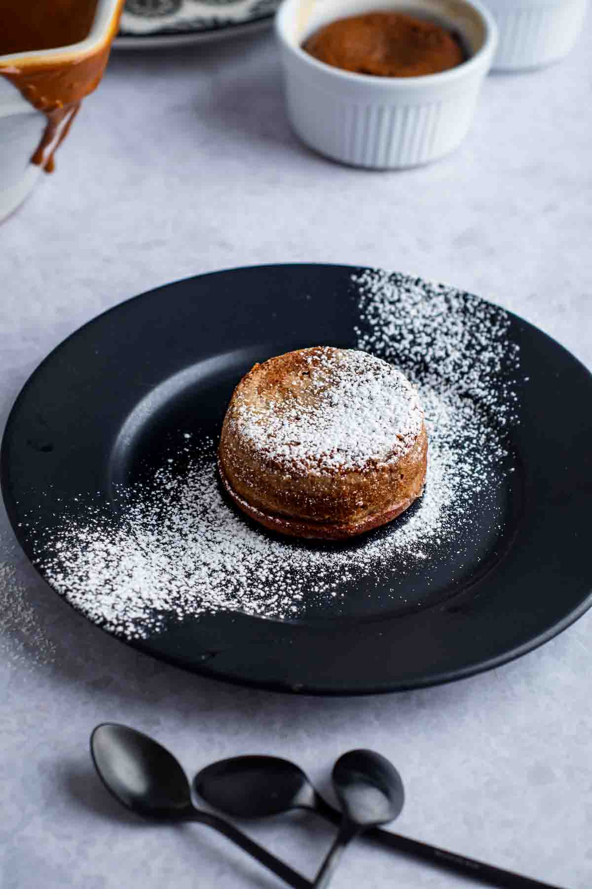 A Dulce de Leche lava cake, decorated with icing sugar, on a black plate, waiting to be cut into.